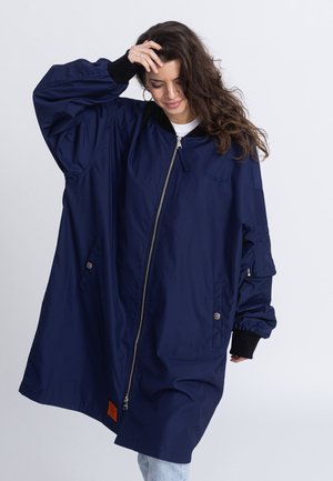 Granatowa, oversize kurtka z zamkiem błyskawicznym z przodu, prążkowanymi czarnymi mankietami, dwoma bocznymi kieszeniami oraz pomarańczową metką na dolnym brzegu. Gładka tekstura.