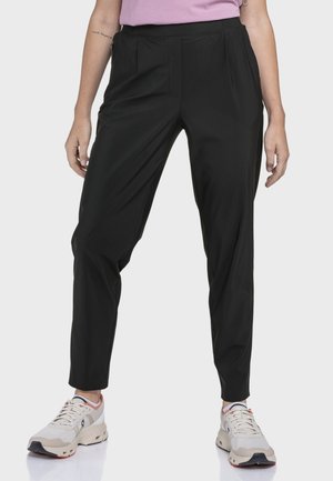 Person trägt schwarze, schmal zulaufende Hose, hellrosa Oberteil und weiße Turnschuhe, steht vor einem schlichten hellen Hintergrund.