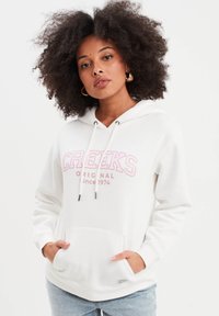 Sudadera blanca con el texto rosa "CREEKS ORIGINAL desde 1974". Presenta un bolsillo frontal, capucha con cordón y puños acanalados. Textura de tela suave.