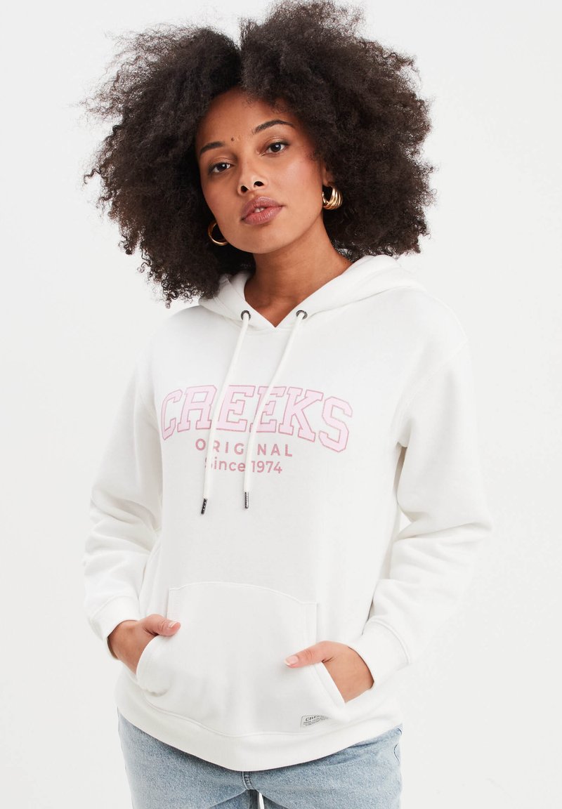 Sudadera blanca con el texto rosa "CREEKS ORIGINAL desde 1974". Presenta un bolsillo frontal, capucha con cordón y puños acanalados. Textura de tela suave.