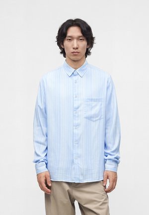 Jeune homme aux cheveux noirs mi-longs porte une chemise à rayures bleues claires à boutons et un pantalon beige, debout devant un fond clair uni.