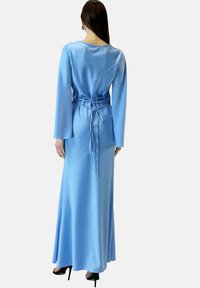 Robe maxi en satin bleu avec des manches longues, un dos nu et un lien en tissu à la taille. Sa texture lisse et son design drapé accentuent l'élégance.