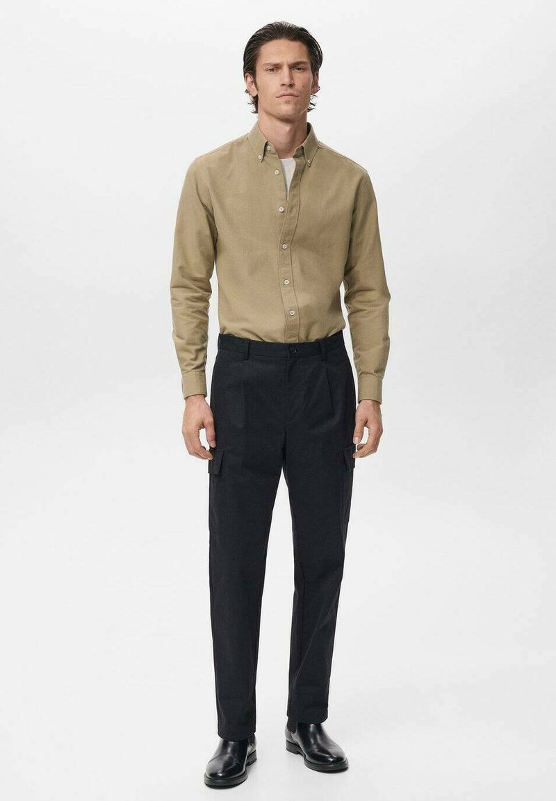Khakifarbenes Hemd mit Knöpfen, schwarze Cargo-Hosen mit Seitentaschen und schwarze Knöchelstiefel, stehend vor einem schlichten weißen Hintergrund.