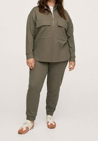 Sudadera verde oliva con dos bolsillos en el pecho y media cremallera, combinada con pantalones joggers a juego. Acentuada con zapatillas de color claro.