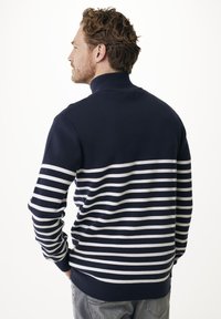 Uomo con capelli ricci che indossa un maglione blu navy con strisce orizzontali bianche, in piedi con le mani in tasca, rivolto a sinistra su uno sfondo bianco.