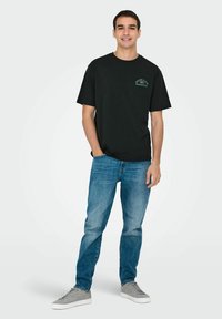 Zwart katoenen t-shirt met ronde hals en korte mouwen, met een groene grafische accenten op de borst, gecombineerd met lichtblauwe jeans en grijze schoenen.