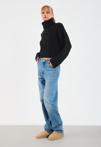 Maglione a collo alto black in maglia a trecce abbinato a jeans dritti blu chiaro e scarpe slip-on in suede beige, ambientato su uno sfondo bianco.