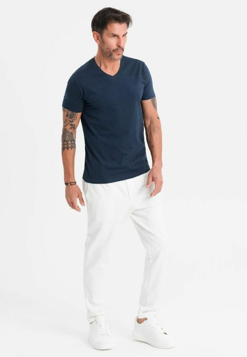 Maglietta navy blue a scollo a V abbinata a pantaloni bianchi a sigaretta e sneakers bianche. La maglietta presenta una texture in cotone liscia, mentre i pantaloni hanno polsini elasticizzati.