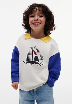 Barn med krøllet hår, smilende, iført en hvid sweatshirt med blå ærmer, gul krave og et sejlskibs motiv, kombineret med blå jeans.