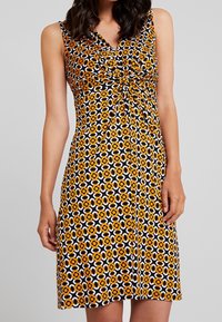 Vestido sin mangas con diseño de escote en V, que presenta un patrón floral en negro, naranja y blanco. Tejido suave con un detalle anudado en la parte delantera.