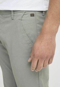 Lichtgrijze katoenen broek met een gestructureerd oppervlak, voorzien van een voorzak en een metalen merklabel op de tailleband. Hand zichtbaar aan de zijkant.