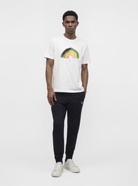 Homme debout portant un t-shirt blanc avec un motif arc-en-ciel multicolore et un pantalon de jogging noir, baskets blanches, sur un fond uni clair.