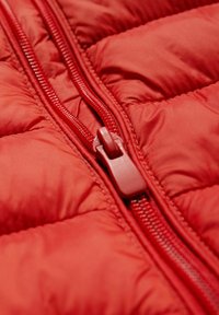 Matériau de veste matelassée rouge avec texture matelassée ; fermeture éclair apparente de couleur assortie ; tirette de fermeture éclair en plastique avec un design circulaire.