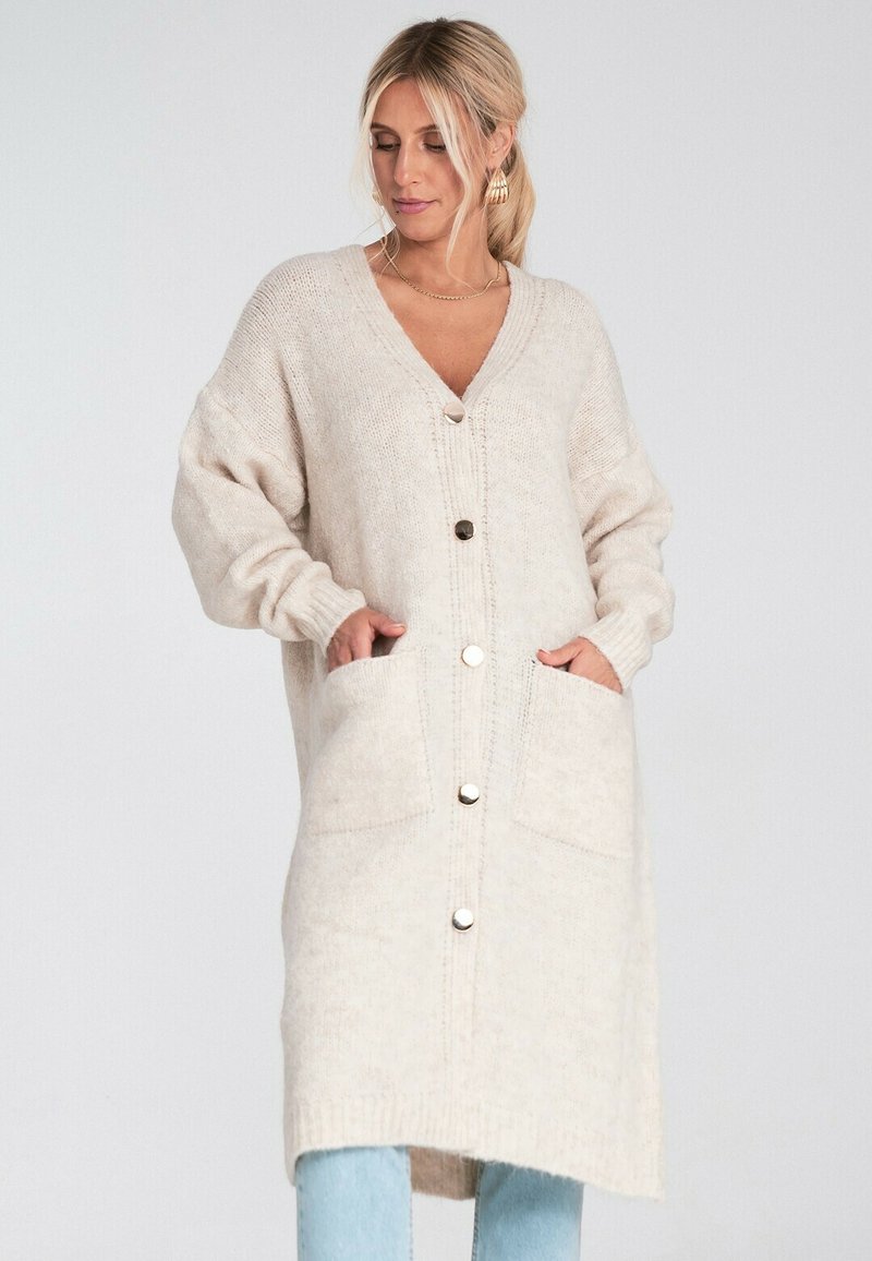 Kobieta w długim beżowym kardiganie zapinanym na guziki, z kieszeniami z przodu, pozująca na tle jednolitym.