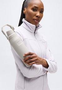 Γυναίκα που φοράει φωτεινό μωβ φλις μπουφάν κρατώντας μια μπεζ μονωμένη παγούρι νερού σε απλό λευκό φόντο.