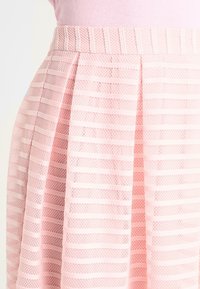 Falda plisada rosa claro con una superposición de malla a rayas horizontales, llevada sobre una falda interior y cinturilla sólidas de color rosa.