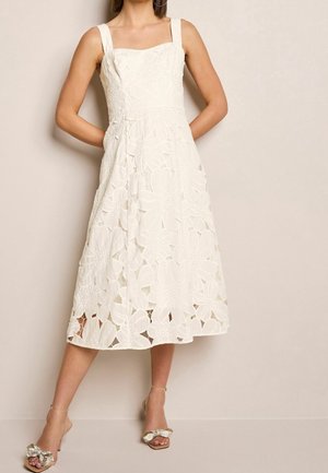 Femme portant une robe midi blanche sans manches avec des motifs floraux découpés et des talons ouverts dorés avec des détails en forme de nœud.