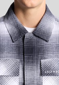 Camicia a quadri con motivi sovrapposti in bianco e nero, colletto rigido, doppie tasche sul petto e un sottile dettaglio del marchio su una tasca.