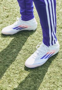 Person som bär vita skor med lila och orangea ränder och lila träningsbyxor som står på konstgräs.