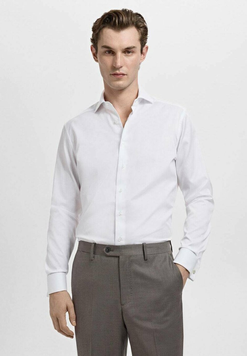 Chemise blanche à col classique, devant boutonné et manches longues, associée à un pantalon gris, avec un tissu lisse et une coupe ajustée.