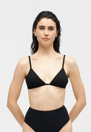 Sötét, nedves hajú nő fekete háromszög bikini felsővel és magas derekú bikini alsóval, egyszerű világos háttér előtt állva.