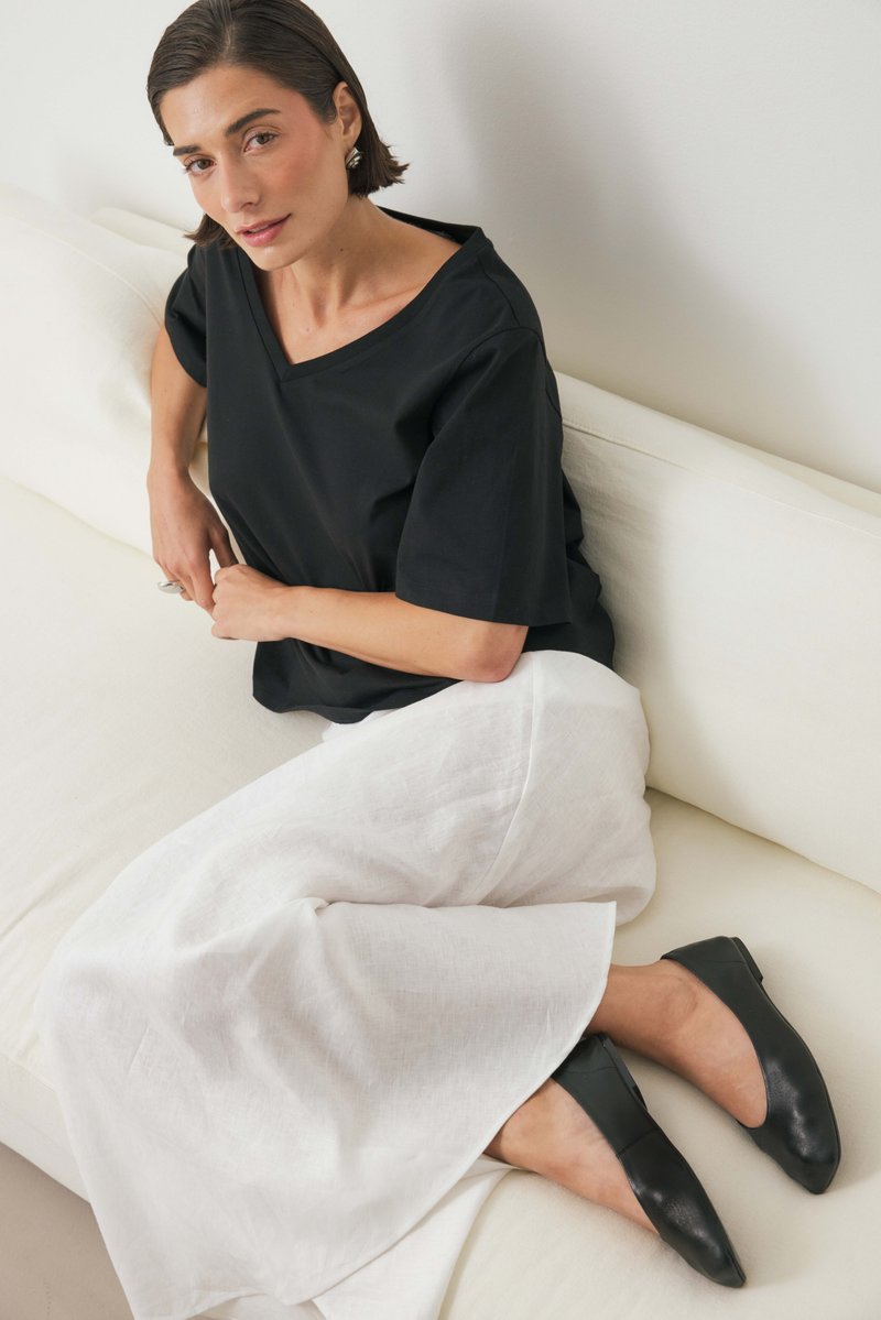 Donna con capelli corti e scuri seduta su un divano bianco, indossa un top nero, una gonna bianca, ballerine nere, orecchini d'argento e un anello.