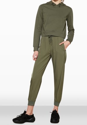Frau trägt olivgrünes Cropped-Hoodie und passende Jogginghose mit schwarzen Sneakern, steht mit einer Hand in der Tasche.