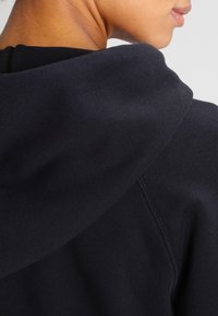 Tmavě námořnická mikina s kapucí s žebrovaným límcem a texturovanou látkou, vybavená kapucí se stahovací šňůrkou a detaily na švech ramen. Hladký vzhled.