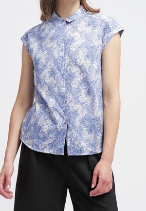 Femme portant une chemise boutonnée à manches courtes, à motifs bleus et blancs, associée à un pantalon noir ample, debout devant un fond uni.