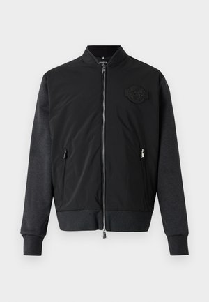 Giubbotto bomber nero con chiusura a zip, polsini a costine e un design in tessuto testurizzato. Presenta una patch con logo sul lato sinistro del petto.
