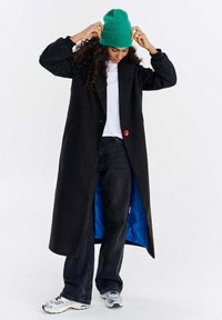 Kobieta poprawiająca zieloną czapkę, ubrana w długi czarny płaszcz z niebieskim podszyciem, białą koszulę, czarne jeansy i białe trampki, stojąca na białym tle.