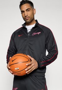 Jachetă cu fermoar Nike neagră, cu accente roșii și textul "Chicago", asociată cu o minge de baschet. Textură netedă, croială sport.