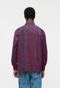 Άνδρας που φορά μακρυμάνικο πουκάμισο με κατακόρυφες κόκκινες και μπλε ρίγες, συνδυασμένο με μπλε τζιν με διακριτικό λουλουδάτο μοτίβο, στέκεται με την πλάτη γυρισμένη.