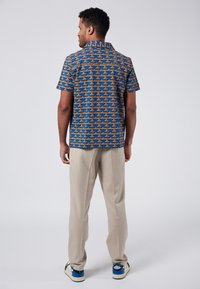 Camisa de manga corta azul con un patrón geométrico en amarillo y crema, combinada con pantalones beige claros y zapatos con detalles azules.