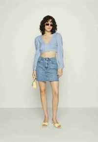 Blusa curta azul clara com mangas bufantes, combinada com uma saia de ganga ajustada. Acessórios incluem sandálias amarelas e uma pequena mala.
