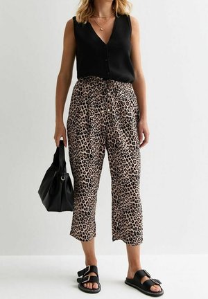 Frau trägt ärmelloses schwarzes Oberteil mit Knöpfen, kurze Leopardenmuster-Hose, schwarze Sandalen und hält eine schwarze Handtasche.