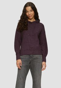 Lila, weich strukturierte Kapuzenjacke mit einer vorderen Tasche und verstellbaren Kordeln. Kombiniert mit grauen Jeans.