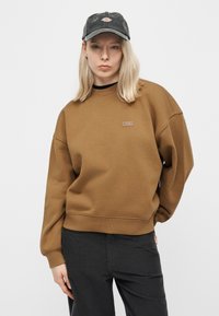 Νεαρή γυναίκα που φοράει καφέ φούτερ και γκρι καπέλο μπέιζμπολ, στέκεται με το ένα χέρι πίσω από την πλάτη της σε απλό φωτεινό φόντο.