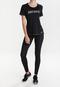 Svart sports t-shirt med texten "JUST DO IT." i silver, tillsammans med svarta leggings med nätpaneler och svarta sneakers.