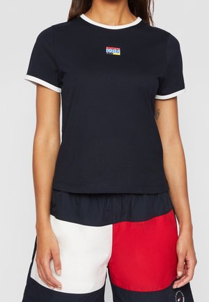 Nainen, jolla on päällään tummansininen t-paita valkoisilla yksityiskohdilla ja pienellä monivärisellä logolla, yhdistettynä värilohkoilla varustettuihin shortseihin, jotka ovat tummansiniset, punaiset ja valkoiset.