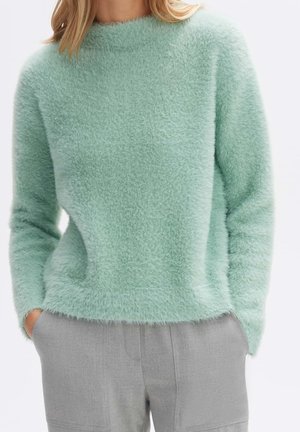 Femme portant un pull vert clair doux et duveteux à manches longues et un pantalon gris, les mains dans les poches.