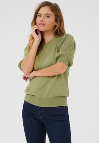Maglione a maglia verde chiaro con maniche corte a sbuffo, scollatura a V e orlo inferiore aderente, abbinato a jeans blu scuro.