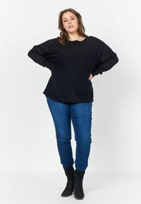 Pullover nero oversize con scollo rotondo e maniche lunghe, abbinato a jeans blu attillati e stivali neri alla caviglia.