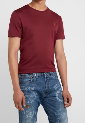 Mężczyzna w prostym, ciemnoczerwonym t-shircie z krótkim rękawem i niebieskich dżinsach z białym graficznym napisem i małą naszywką tygrysa na lewym udzie.