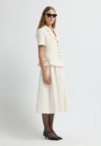 Donna che indossa un blazer corto crema con bottoni, una gonna midi plissettata abbinata, collant neri, décolleté neri con tacco a kitten e occhiali da sole a occhio di gatto.