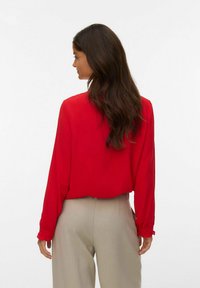 Mujer con cabello largo y oscuro, vistiendo una blusa roja de manga larga y pantalones beige, de espaldas frente a un fondo blanco liso.