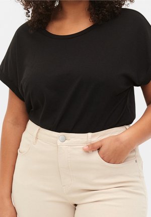 Kobieta ubrana w czarną krótka koszulkę z krótkim rękawem, wpuszczoną w wysokowodne beżowe spodnie, z prawą ręką w kieszeni.