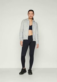 Sudadera gris con cremallera, sujetador deportivo negro, leggins negros, zapatillas negras. La modelo está de pie de frente contra un fondo neutro.