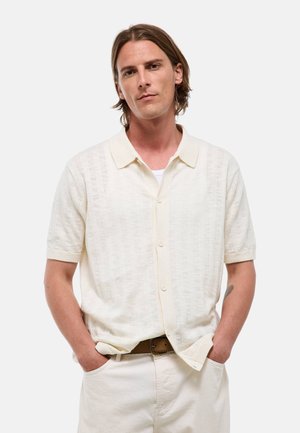 Homme aux cheveux mi-longs portant une chemise crème à manches courtes boutonnée et un pantalon crème, les mains dans les poches sur fond blanc.