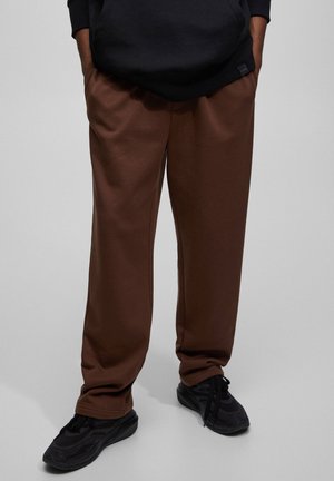 Unterkörper trägt braune locker sitzende Hose, schwarze Sneakers und einen schwarzen Hoodie mit Händen in den Taschen vor einfarbigem Hintergrund.